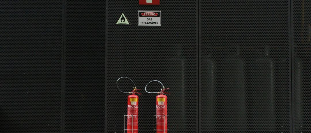 Zwei rote Feuerlöscher stehen auf einem Ständer vor einer schwarzen Netzwand. Über den Feuerlöschern befinden sich Schilder, die vor brennbarem Gas warnen.