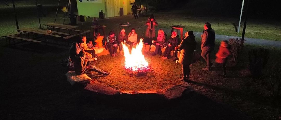 Menschen versammeln sich abends um ein Lagerfeuer, einige auf Bänken sitzend und andere stehend. Ein Gebäude mit einem Schild, das 'Spielplatz und Parkanlage Altenberg' besagt, ist im Hintergrund sichtbar.