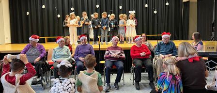 Eine Gruppe von Kindern in weißen Kostümen, die auf der Bühne stehen und vor einem Publikum auftreten. Erwachsene sitzen in der ersten Reihe, mit Weihnachtsmützen. Schneeflocken hängen am Bühnenhintergrund.