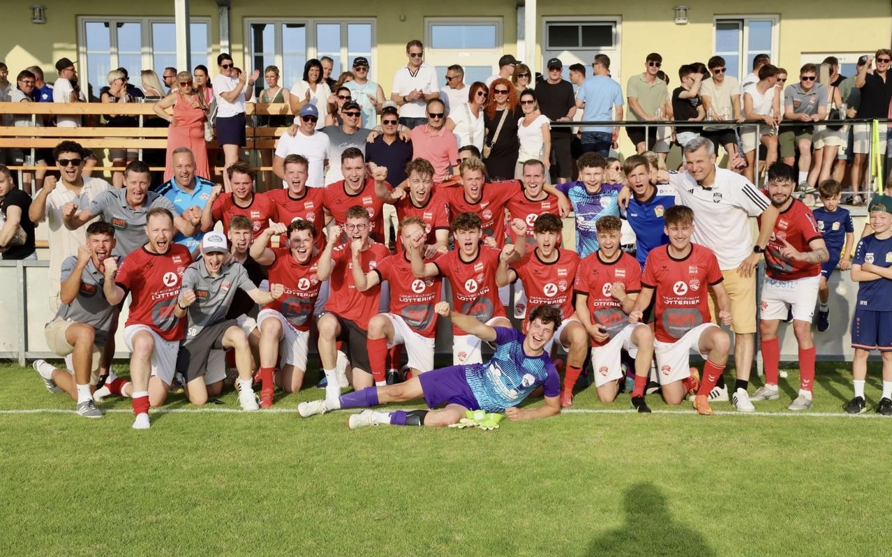 Eine Fußballmannschaft feiert auf einem Spielfeld, während eine Menge hinter einem Zaun zuschaut. Die Spieler tragen rote und weiße Trikots mit Lotto-Sponsoren.