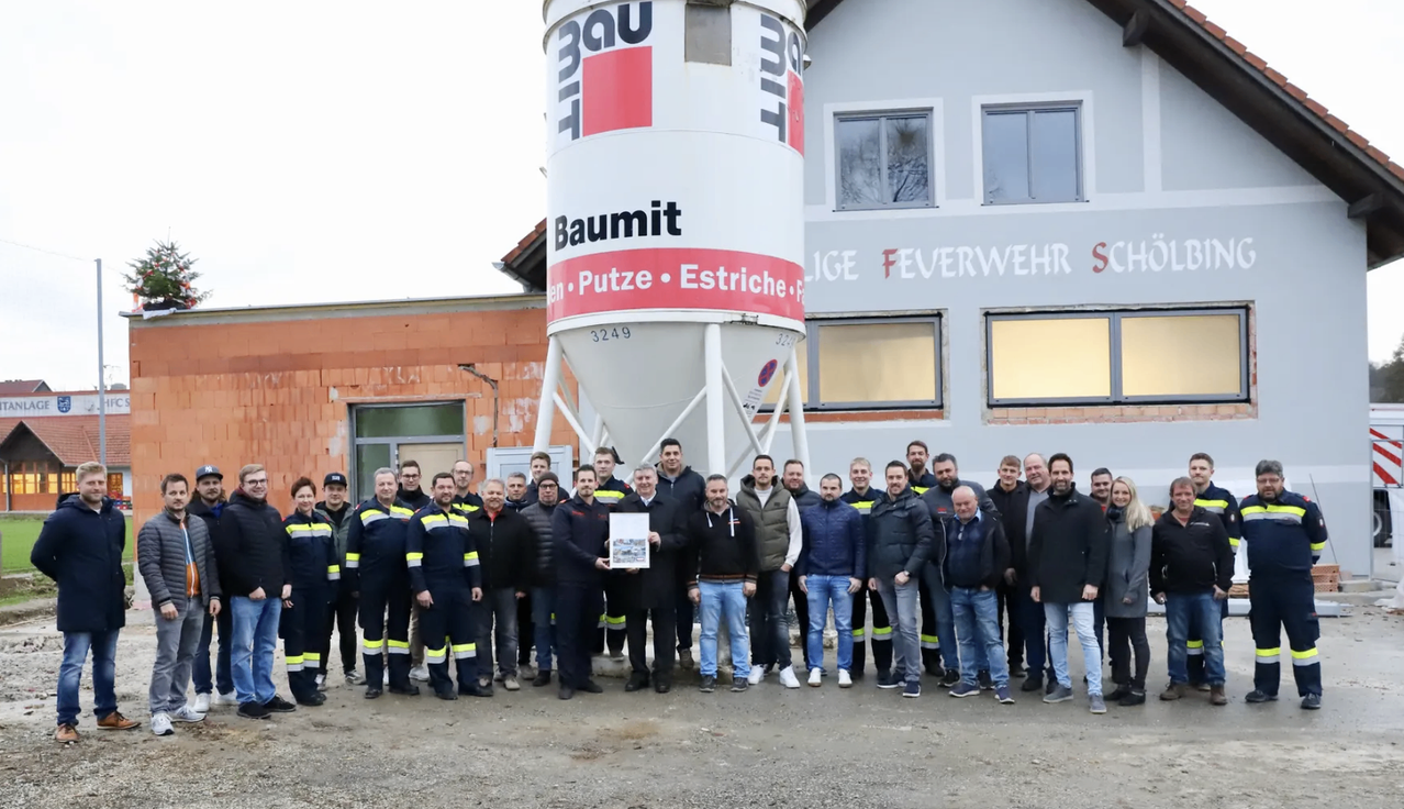 Eine Gruppe von Feuerwehrleuten in Uniformen steht vor einem Baumit-Gebäude und hält ein gerahmtes Zertifikat. Das Gebäude hat einen großen zylindrischen Lagertank mit Baumit-Marke.