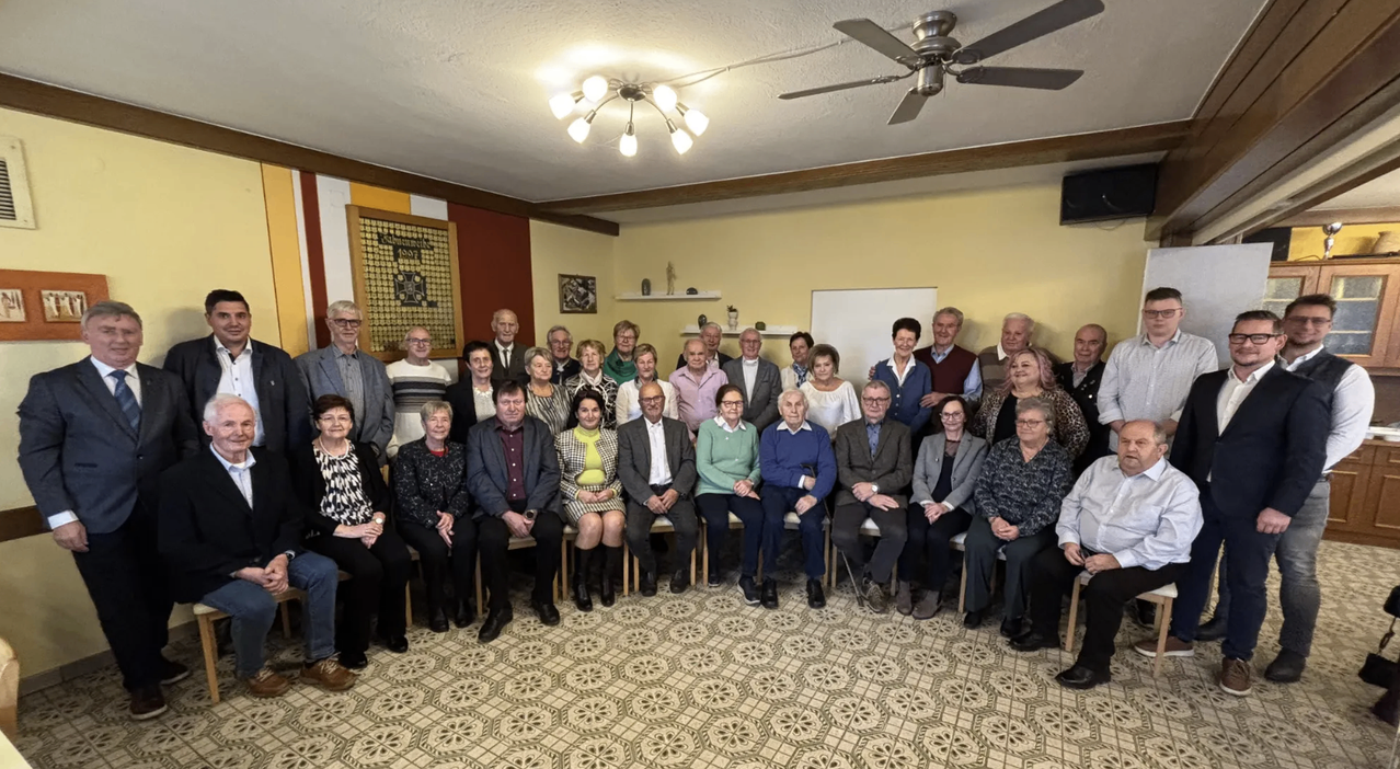 Eine Gruppe von Menschen, möglicherweise bei einem Wiedersehen, posiert für ein Foto in einem Raum mit gemustertem Boden, gelben Wänden und verschiedenen Dekorationen. Eine Deckenventilator und eine Tafel sind ebenfalls zu sehen.