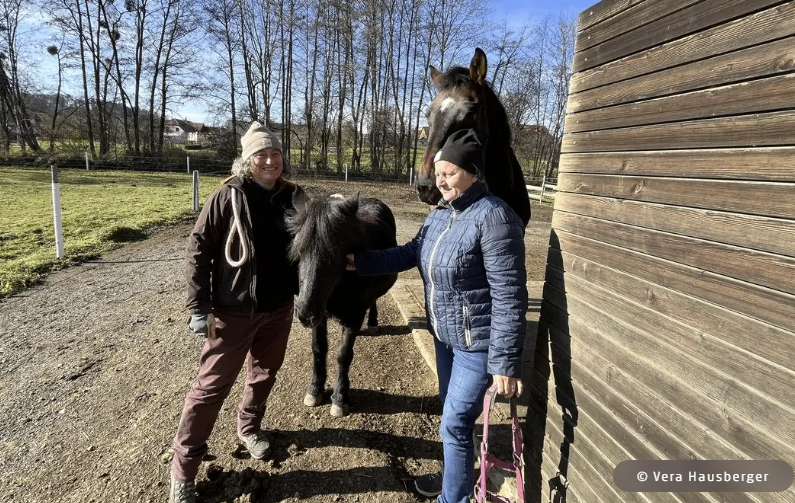 Zwei Frauen stehen neben einem Pferd und lächeln. Ein größeres Pferd steht hinter ihnen. Sie befinden sich in der Nähe einer Holzwand. Ein Feld und Bäume sind im Hintergrund zu sehen.