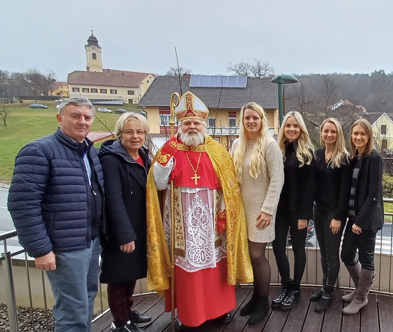 Eine Gruppe von Menschen posiert für ein Foto auf einer Terrasse, mit einer Kirche im Hintergrund. Ein älterer Mann und eine Frau stehen links, während ein Mann in geistlicher Kleidung in der Mitte steht. Drei Frauen stehen rechts, alle lächeln.