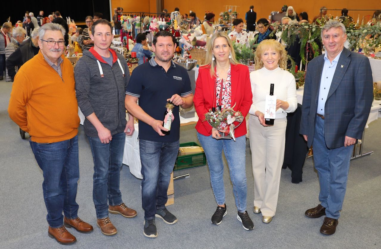 Fünf Menschen stehen in einem Raum eng beieinander. Drei Männer und zwei Frauen halten eine Flasche Wein. Sie alle lächeln. Hinter ihnen stehen Tische mit vielen ausgestellten Gegenständen.