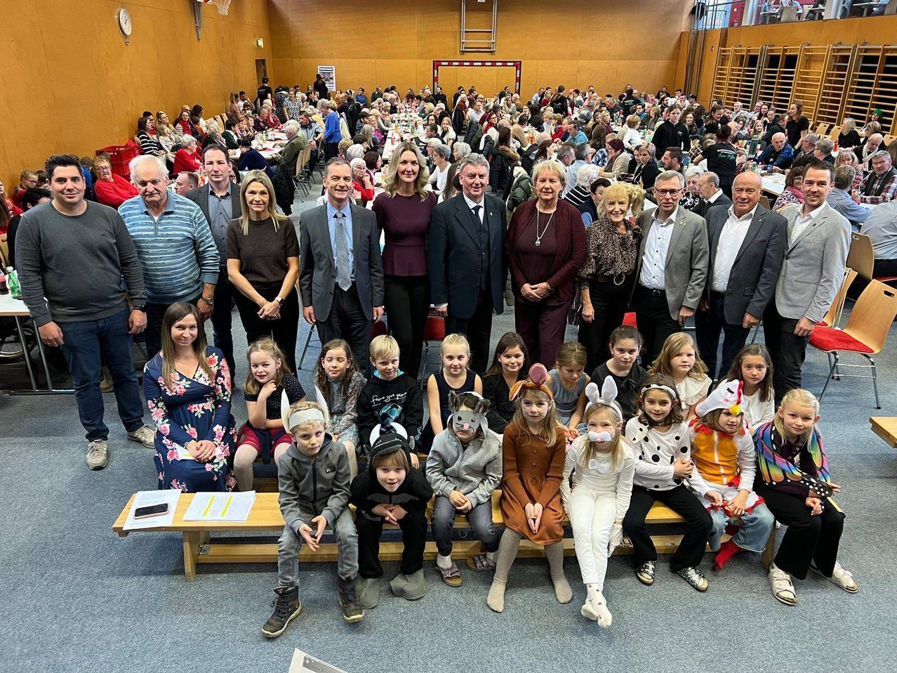 Eine große Gruppe von Menschen, darunter Erwachsene und Kinder, versammelt sich in einem Saal. Einige sind in Kostümen gekleidet. Sie posieren für ein Foto vor einer Menschenmenge.