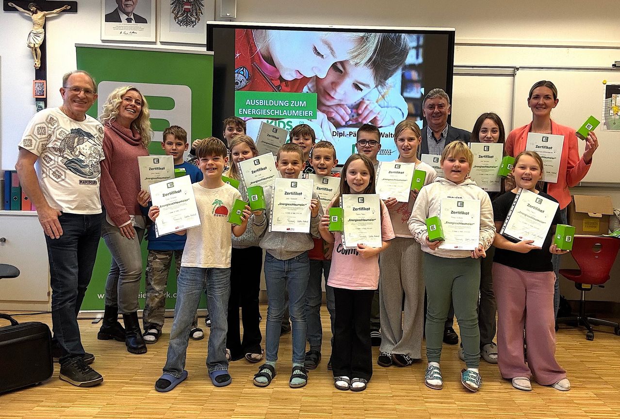 Kinder und Erwachsene halten Zertifikate in der Hand und stehen in einer Reihe in einem Raum. Eine Beamerleinwand zeigt das Gesicht eines Kindes und die Worte 'Ausbildung zum Energieschlaumeier'.