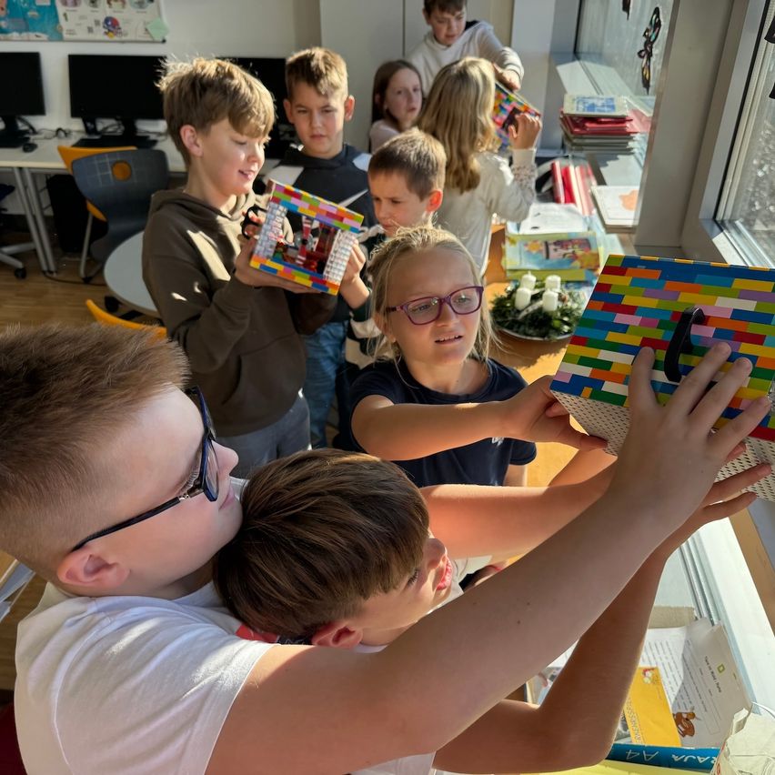 Eine Gruppe von Kindern in einem Klassenzimmer baut mit bunten Legosteinen. Ein Junge hält eine Box, während andere zuschauen oder fertige Strukturen halten.