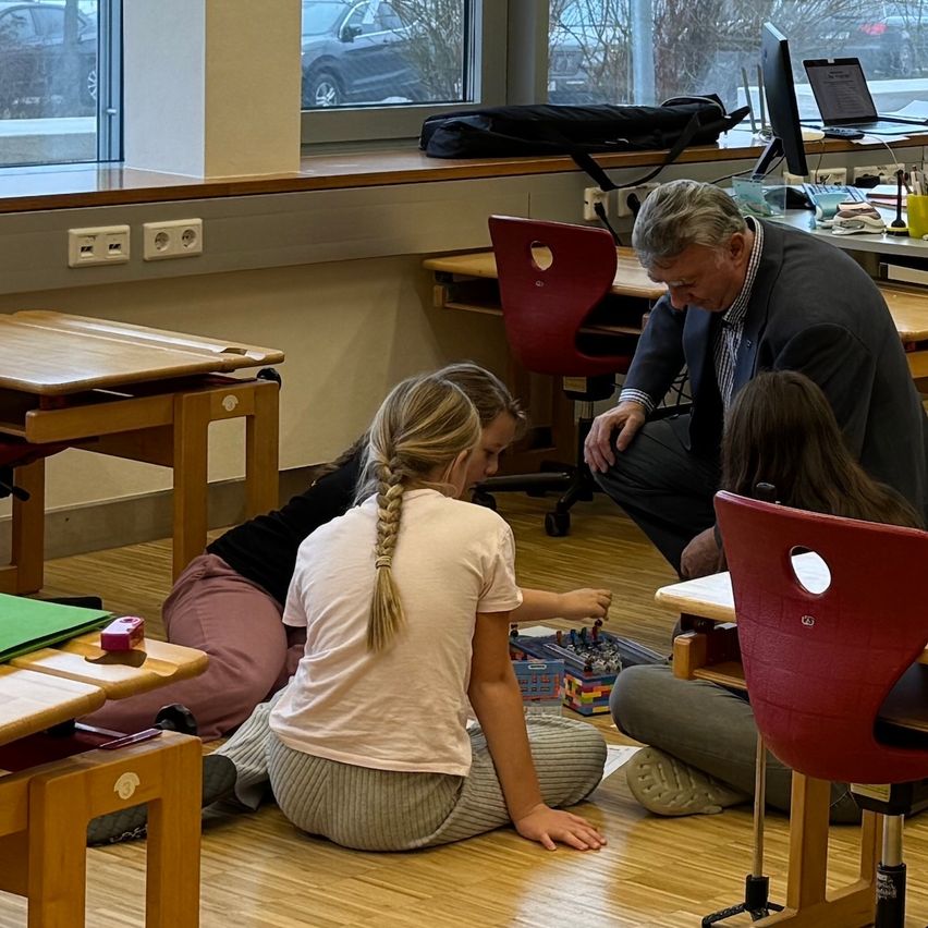 In einem Klassenzimmer hilft ein Mann zwei Mädchen bei einem Lego-Projekt, während ein Auto durch das Fenster zu sehen ist.
