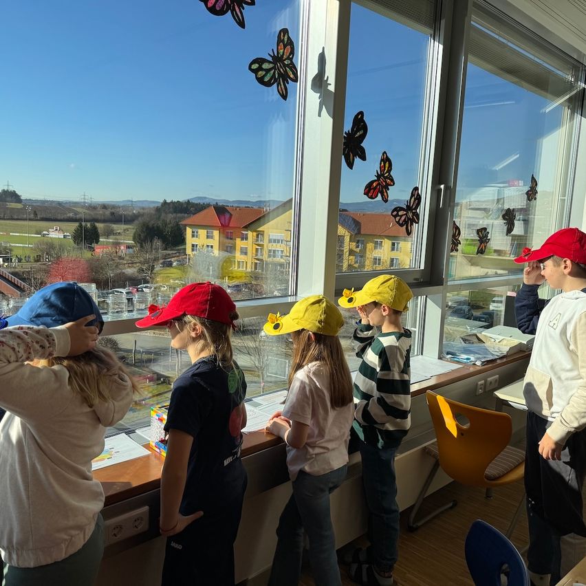 Mehrere Kinder mit Hüten schauen aus einem Fenster mit Schmetterlingsaufklebern. Im Hintergrund ist ein Gebäude sichtbar.