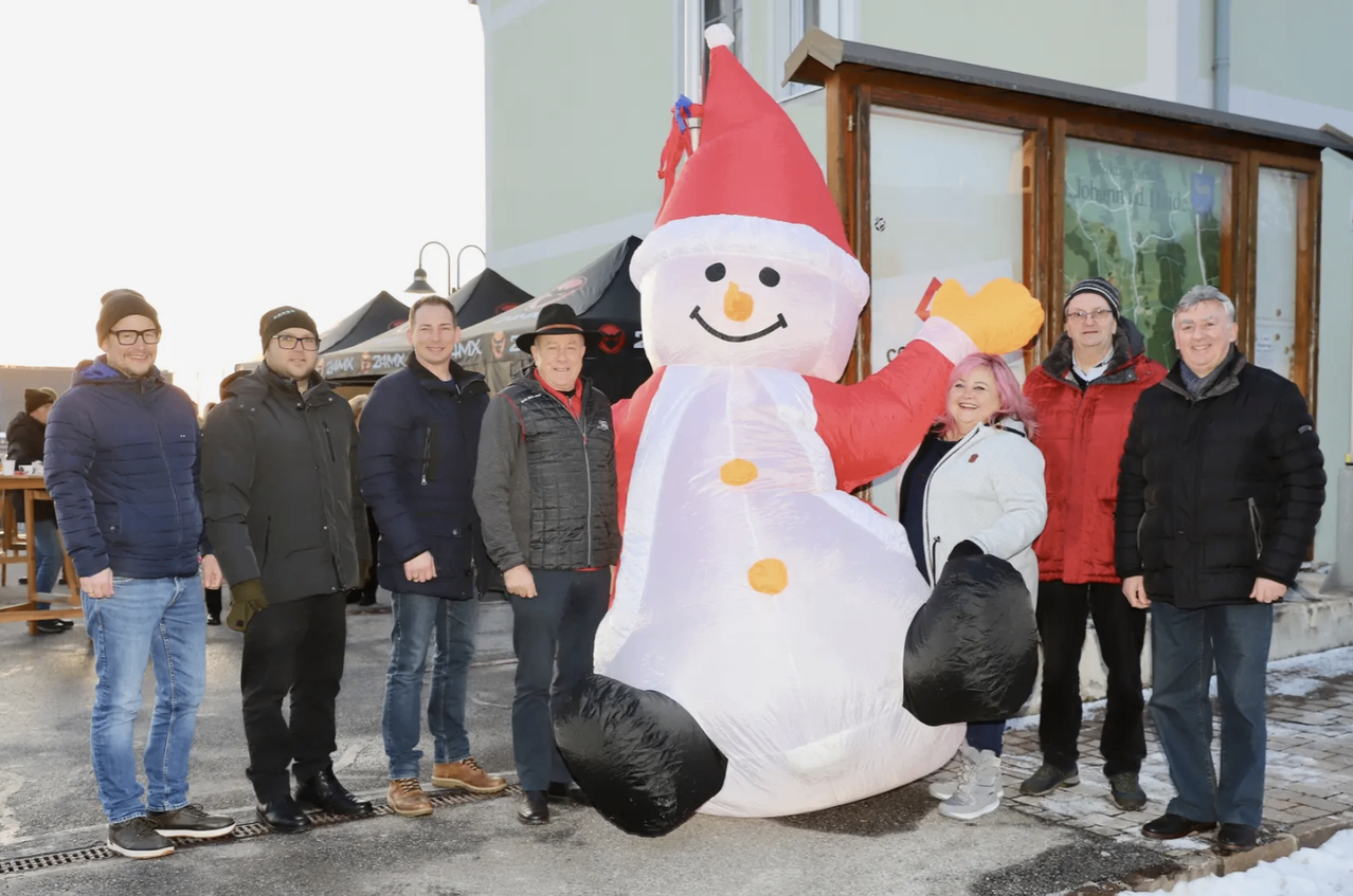 Fünf Personen, darunter zwei Frauen, stehen vor einem riesigen aufblasbaren Schneemann, der einen Weihnachtsmannhut und Handschuhe trägt, in einer winterlichen Umgebung.