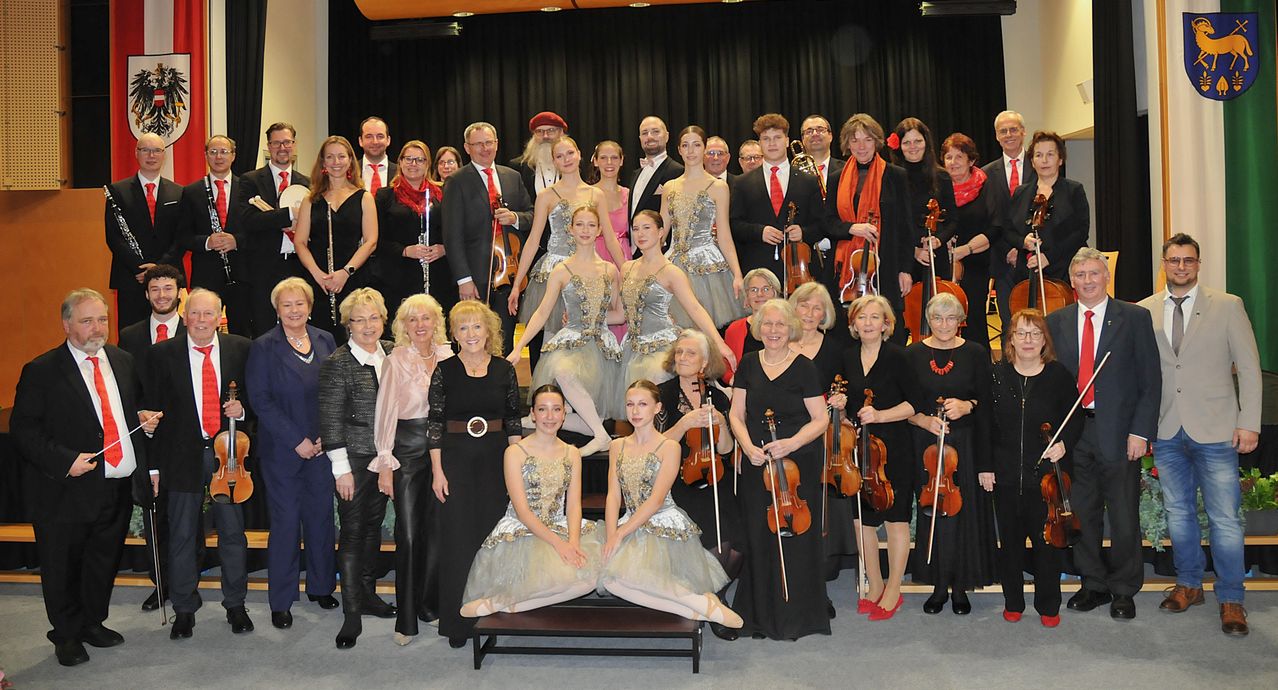 Eine Gruppe von Musikern und Tänzern posiert für ein Foto vor einem schwarzen Hintergrund. Einige tragen elegante Kleider, andere halten Geigen. Sie alle lächeln.