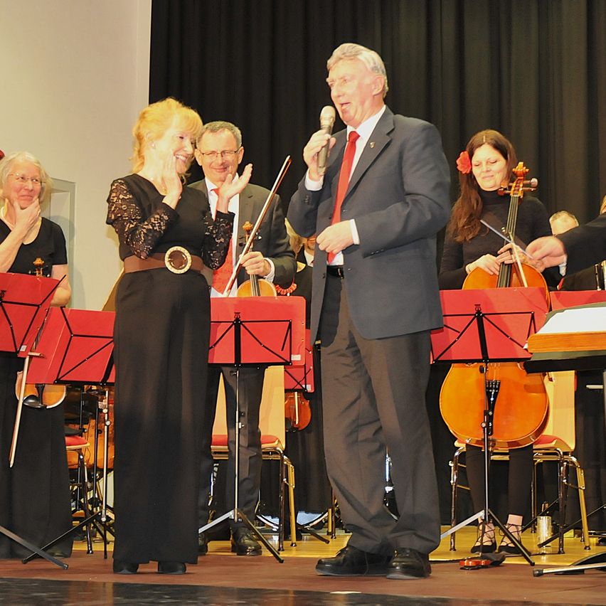 Ein Mann mit Mikrofon spricht zu einer Frau auf der Bühne. Hinter ihnen spielen Musiker Violinen und Celli. Das Publikum schaut zu.