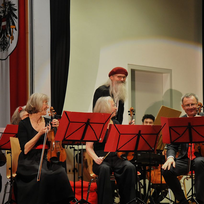 Eine Gruppe von Musikern steht auf der Bühne, darunter eine Violinistin und eine Geigerin mit Bogen. Sie sind alle in Schwarz gekleidet und sitzen auf Stühlen, mit roten Notenständern vor ihnen. Ein Mann steht hinter ihnen und trägt eine Baskenmütze. An der Wand hängen eine Flagge und eine Tür.