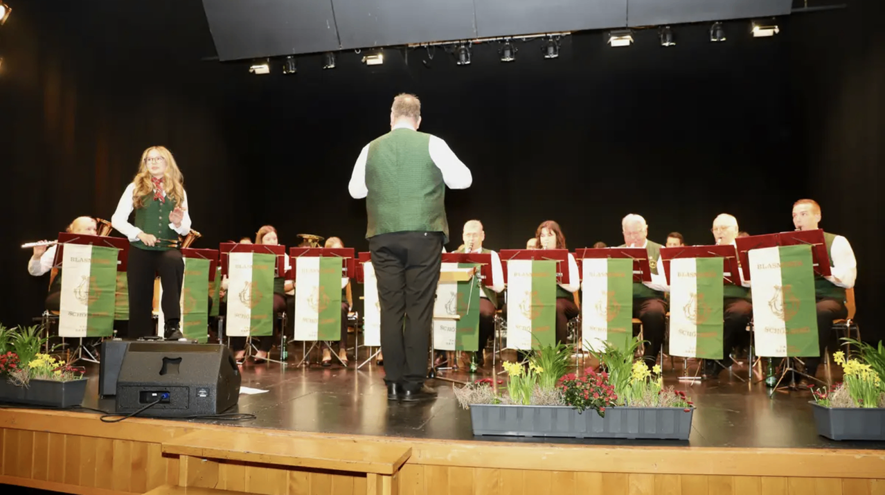 Eine Band steht auf der Bühne, ein Mann vorne. Dahinter sitzen Bandmitglieder in Stühlen mit Notenständern. Die Bühne ist mit grünen Bannern und Blumen dekoriert.