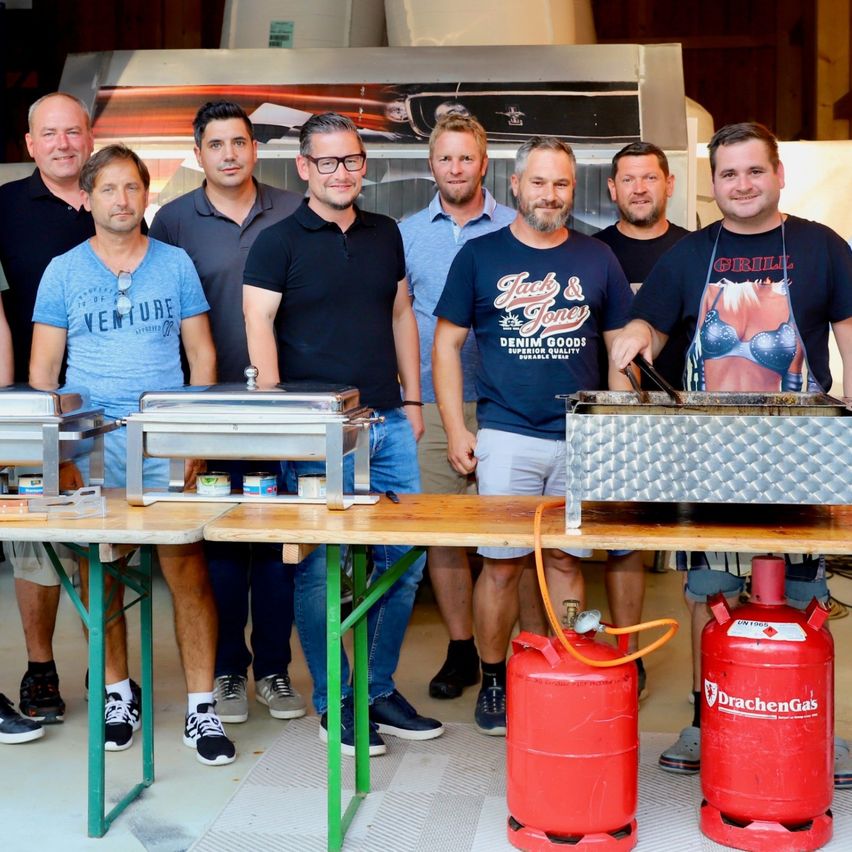 Eine Gruppe von Männern in Freizeitkleidung steht hinter einem Tisch mit Essensschalen und Propangasflaschen und lächelt für ein Foto.