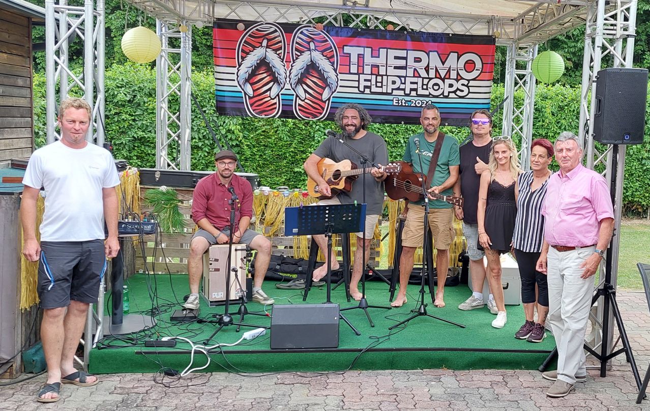 Eine Gruppe von Musikern steht mit Gitarren auf einer Bühne und lächelt für ein Foto. Ein Banner mit dem Text 'THERMO FLIP-FLOPS' hängt hinter ihnen.