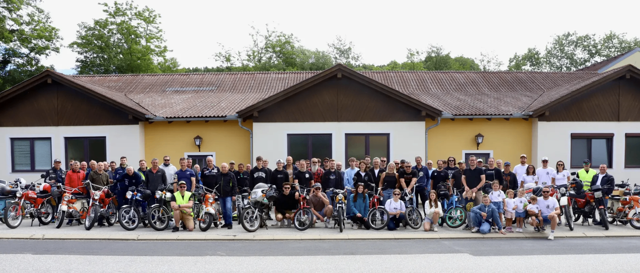 Eine große Gruppe von Menschen posiert für ein Foto vor einem Haus mit ihren Fahrrädern und Motorrädern.