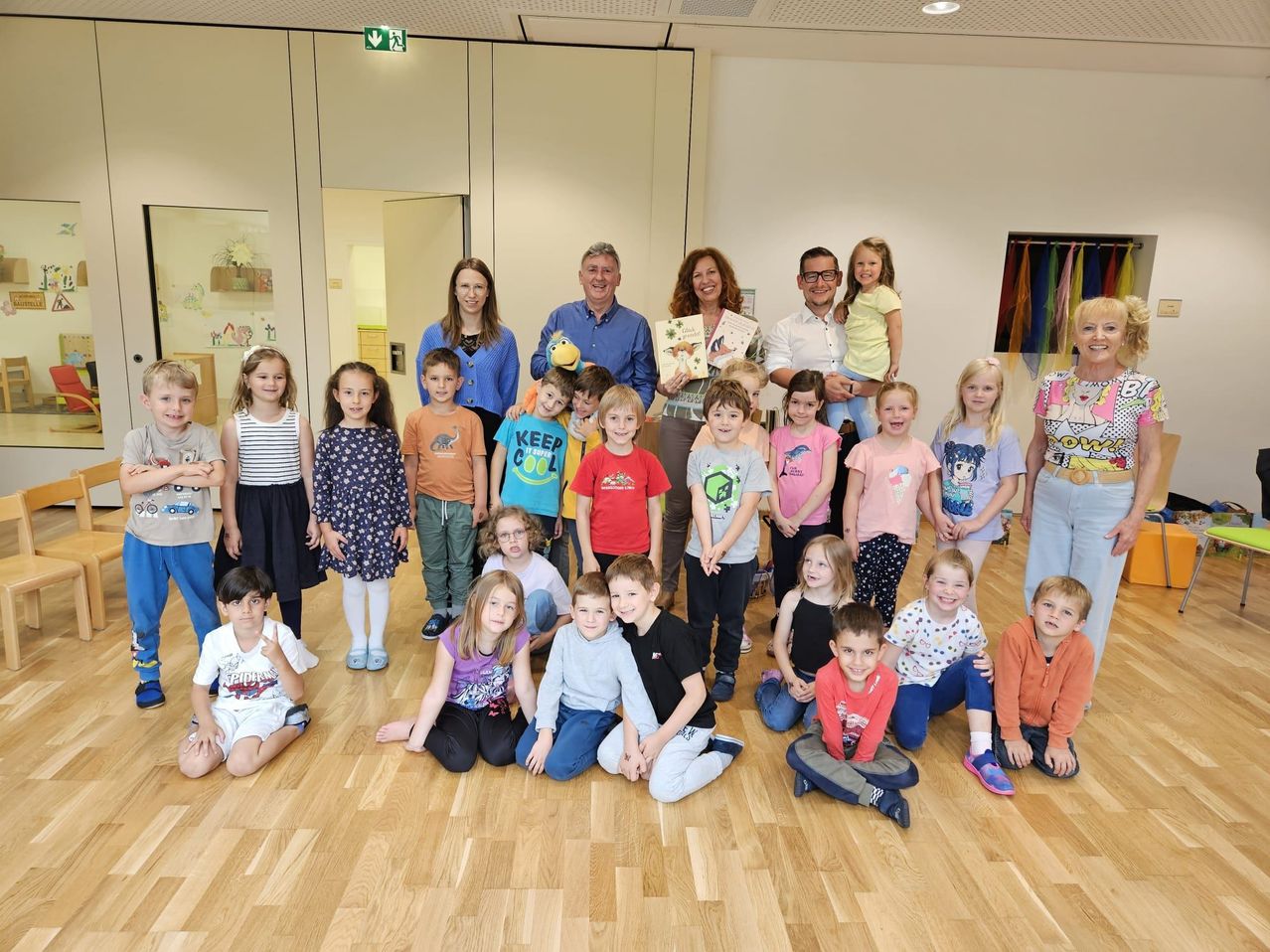 Gruppe von Kindern und Erwachsenen posiert für ein Foto in einem Raum mit Holzböden und Glaswänden, hält Bücher und lächelt.