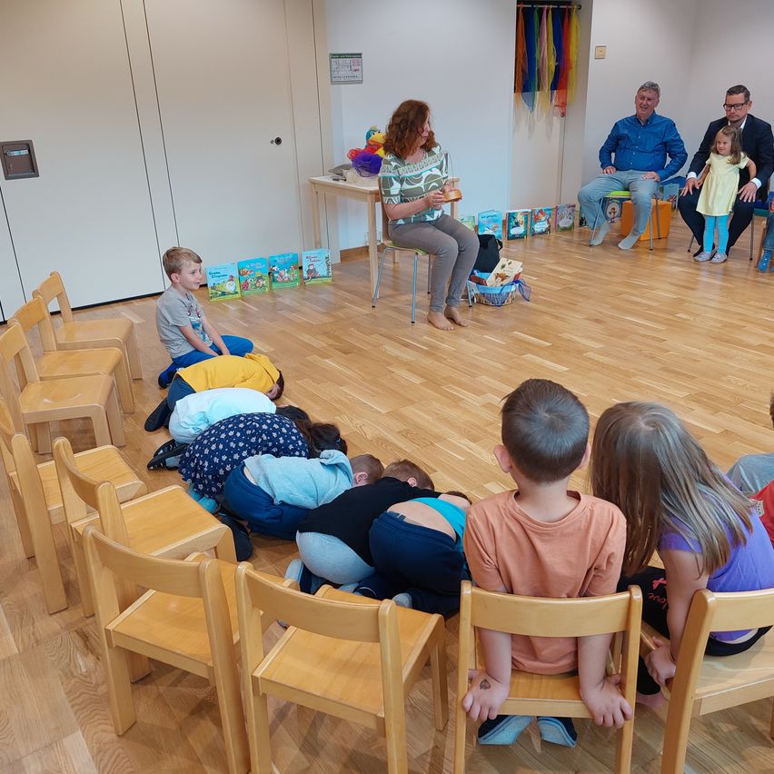 Eine Frau liest Kindern vor, die in einem Kreis sitzen. Erwachsene sitzen im Hintergrund. Bunte Spielzeuge und Bücher liegen auf dem Boden. Der Raum hat Holzböden und weiße Wände.