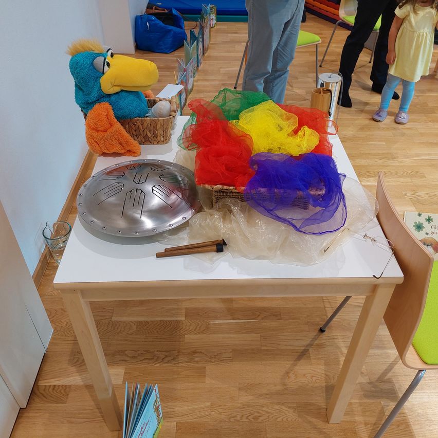 Ein weißer Tisch mit verschiedenen farbigen Gegenständen, einer Metallplatte, einem Korb mit einem Papageienspielzeug und einigen Büchern auf dem Boden. Es stehen Leute um den Tisch herum.