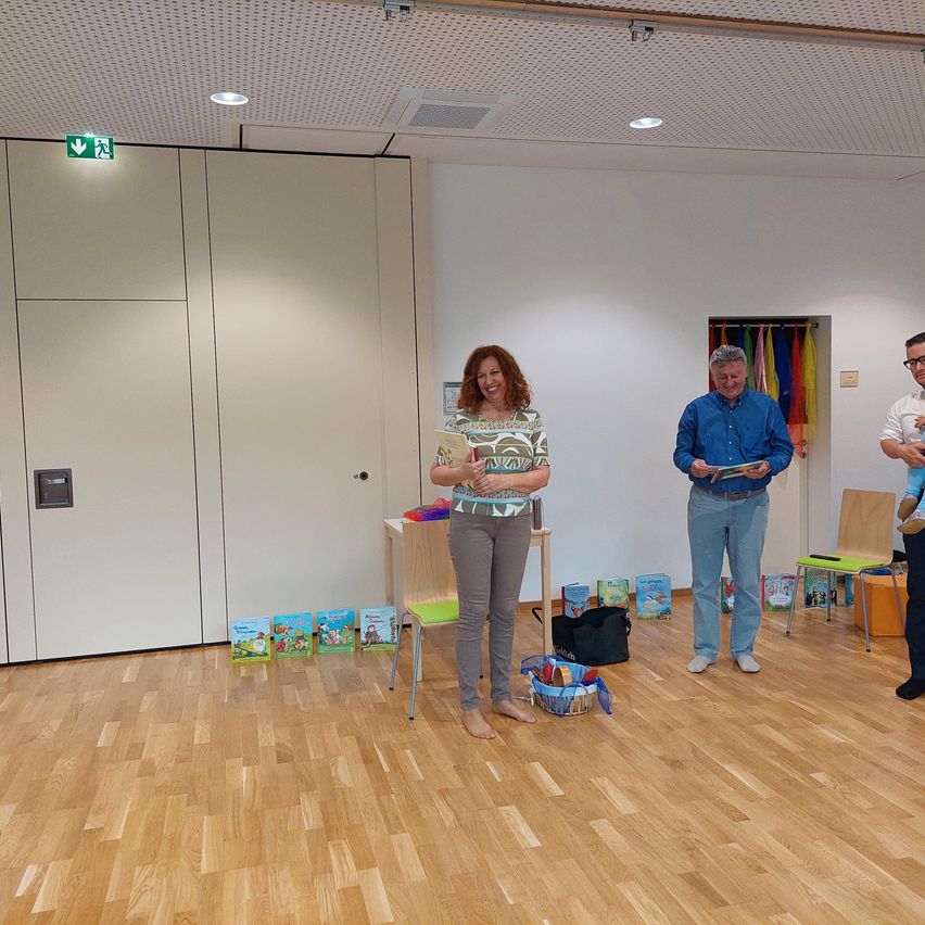Vier Menschen stehen in einem Raum mit Holzböden, eine Frau hält ein Buch, ein Mann liest, ein anderer Mann hält ein Baby, und ein Stuhl mit Büchern ist in der Nähe.