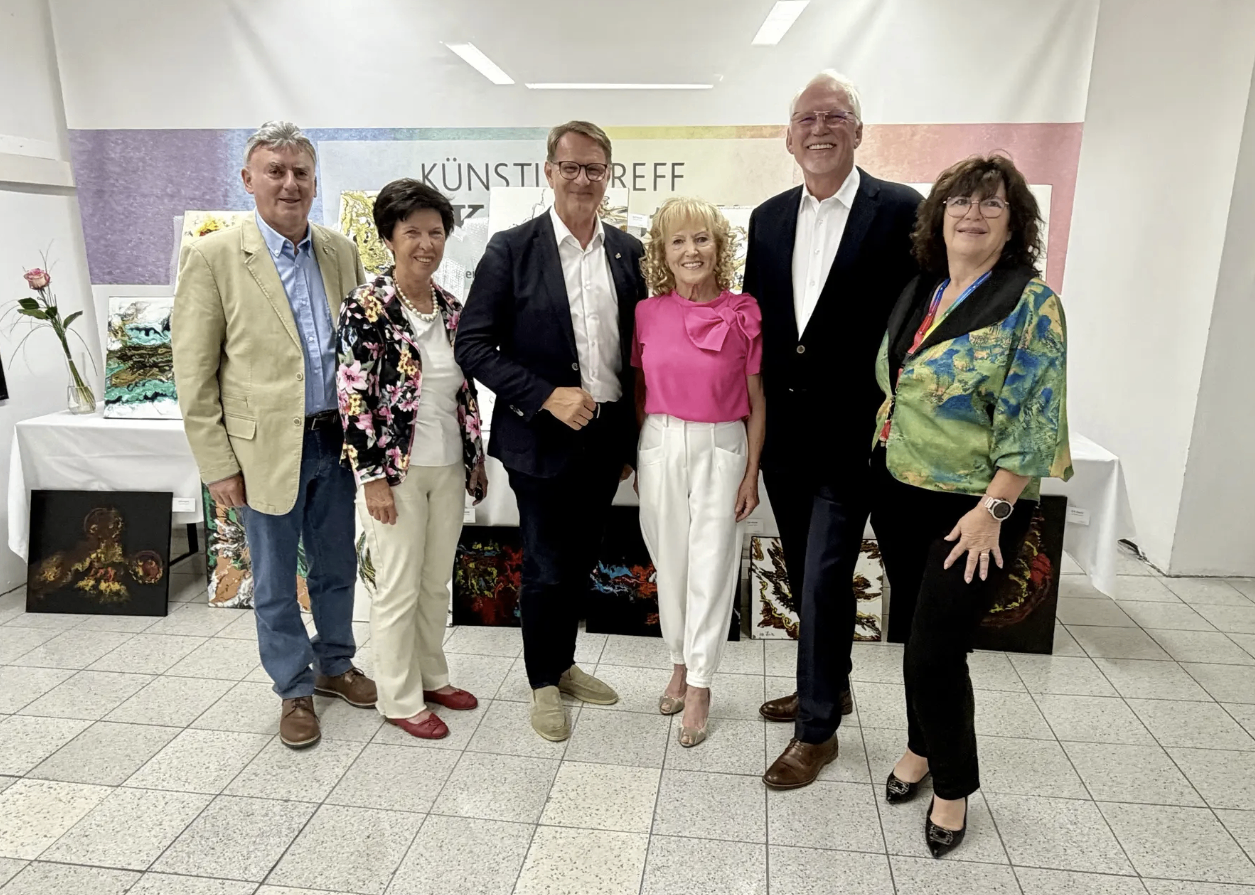 Sechs Personen posieren für ein Foto in einem Raum mit farbenfrohem Hintergrund. Zwei Frauen und vier Männer, alle leger gekleidet. Die Frau in Pink lächelt fröhlich.