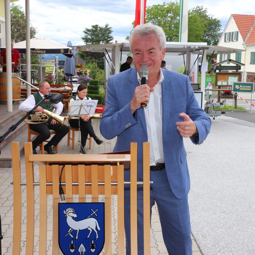 Ein Mann spricht in ein Mikrofon bei einer Outdoor-Veranstaltung. Dahinter spielen Musiker Instrumente. Ein Haus und ein Schild sind im Hintergrund.