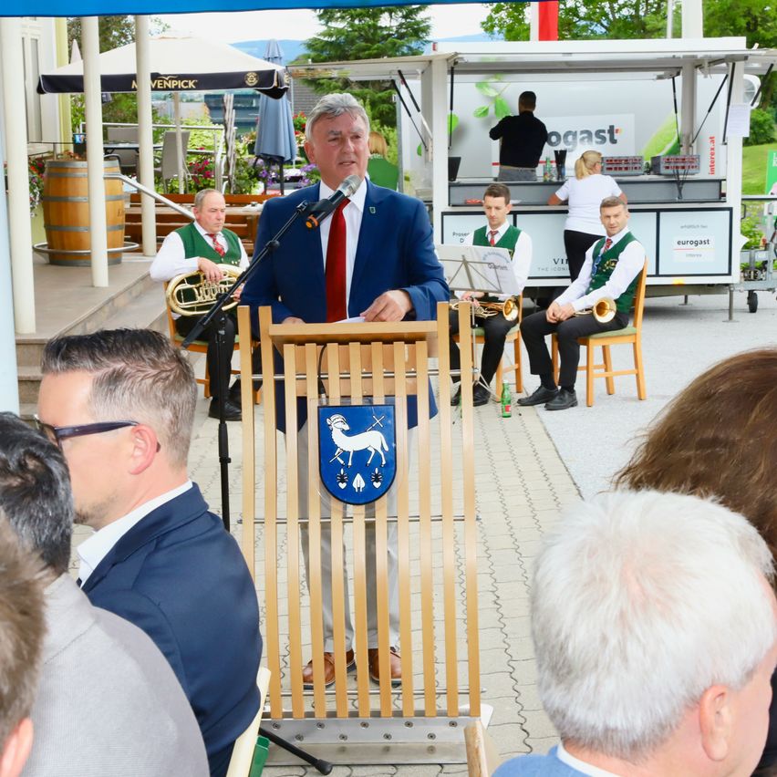 Ein Mann spricht in ein Mikrofon an einem Podium vor einer Menge. Zwei Musiker sitzen hinter ihm und ein Imbisswagen ist im Hintergrund zu sehen.