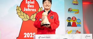 Ein Mann in einem roten Jackett hält eine hölzerne Trophäe mit der Aufschrift Spiel des Jahres 2025 vor einem Hintergrund, der einen roten Kreis mit derselben Aufschrift zeigt.