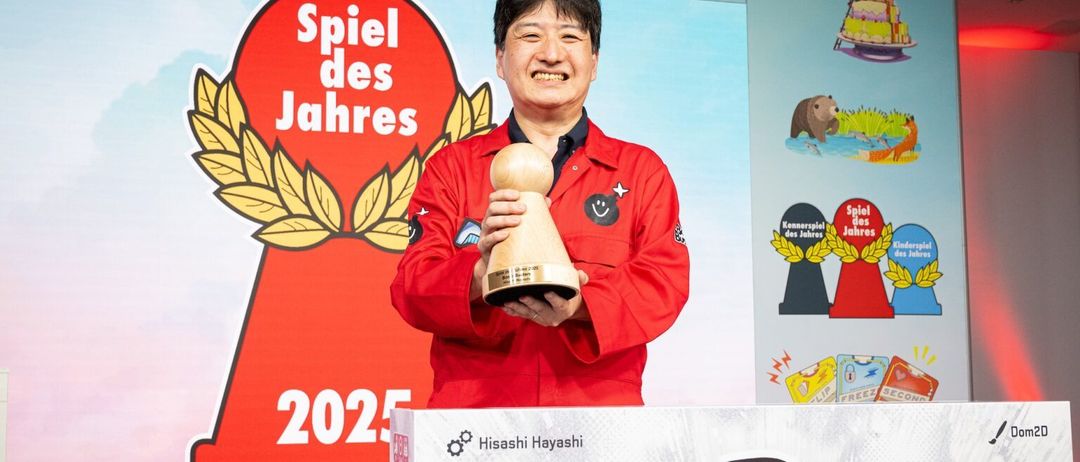 Ein Mann in einem roten Jackett hält eine hölzerne Trophäe mit der Aufschrift Spiel des Jahres 2025 vor einem Hintergrund, der einen roten Kreis mit derselben Aufschrift zeigt.