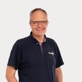 Ein Mann in einem dunkelblauen Poloshirt mit Logo auf der linken Brust, gepaart mit blauer Jeans und schwarzen Sneakern, steht lächelnd mit den Händen in den Taschen.