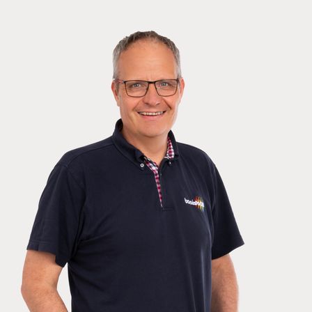 Ein Mann in einem dunkelblauen Poloshirt mit Logo auf der linken Brust, gepaart mit blauer Jeans und schwarzen Sneakern, steht lächelnd mit den Händen in den Taschen.