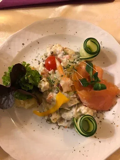Ein Teller mit Kartoffelsalat, garniert mit Gurke, Tomate und Lachs auf einer weißen Tischdecke.