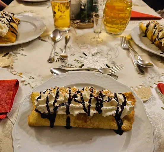 Ein Tisch mit festlicher Einrichtung, auf dem ein Dessert auf einem Teller mit Schlagsahne und Schokoladen-Dreschtupfer steht. Zwei Biergläser und ein kleines Glas sind neben dem Dessert platziert. Der Tisch ist mit einer weißen Tischdecke mit Blumenmuster bedeckt.