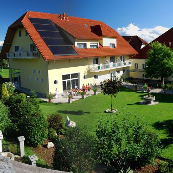 Luftaufnahme eines gelben Hauses mit Solarpanelen auf dem Dach. Menschen sind draußen und genießen den Tag. Das Haus ist von viel Grün umgeben, einschließlich Bäumen und Büschen.