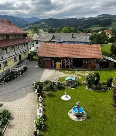 Luftaufnahme eines großen Hauses mit einer Einfahrt und einem kleinen Garten, umgeben von einem grünen Feld mit Bergen im Hintergrund.
