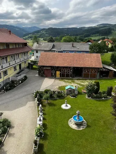 Luftaufnahme eines großen Hauses mit einer Einfahrt und einem kleinen Garten, umgeben von einem grünen Feld mit Bergen im Hintergrund.