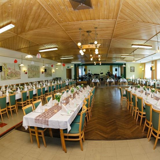 Ein Restaurant mit Holzwänden und -boden, ausgestattet mit langen Tischen, Blumen, Stühlen und Kronleuchtern. In der Ferne ist eine Bühne mit einer stehenden Person zu sehen.