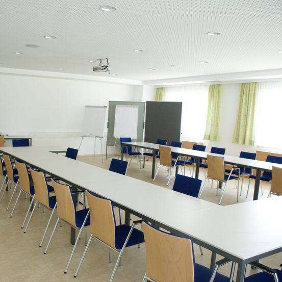Ein leerer Klassenraum mit langen weißen Tischen, blauen Stühlen, weißen Whiteboards und gelben Vorhängen. Die Decke hat versenkte Beleuchtung.