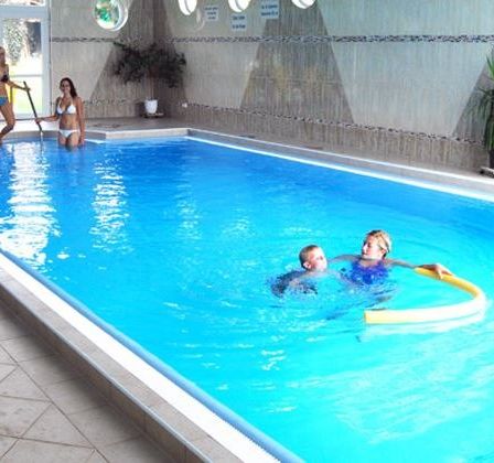Ein rechteckiger Innenpool mit zwei Schwimmenden und zwei Frauen am Rand. Ein Topf mit Pflanzen steht rechts.