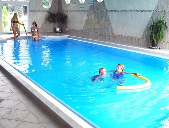Ein rechteckiger Innenpool mit zwei Schwimmenden und zwei Frauen am Rand. Ein Topf mit Pflanzen steht rechts.