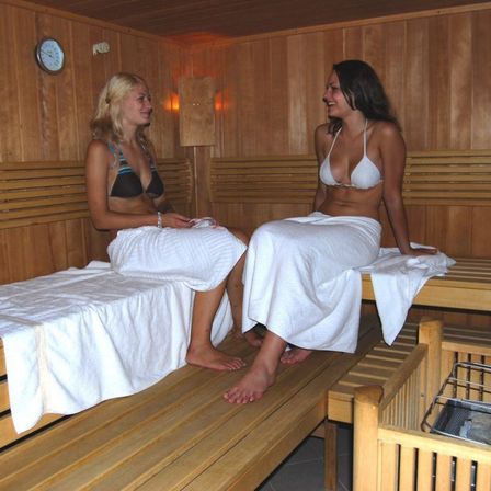 Zwei Frauen in Bikinis sitzen auf Holzbänken in einer Sauna. Eine Frau trägt ein Handtuch um den Hals, und die andere lächelt.