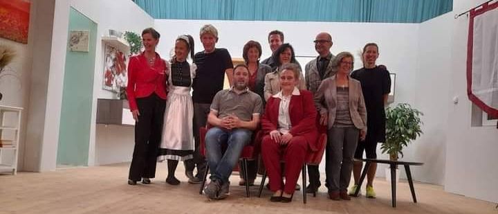 Eine Gruppe von Menschen, möglicherweise ein Theaterensemble, steht und sitzt auf einer Bühne, lächelt und posiert für ein Foto. Die Bühne hat einen Holzboden und einen blauen Vorhang als Hintergrund.