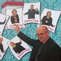 Ein Mann und mehrere Frauen posieren für ein Foto mit dem Wort Samstag darauf. Der Mann lächelt und zeigt auf die Kamera.