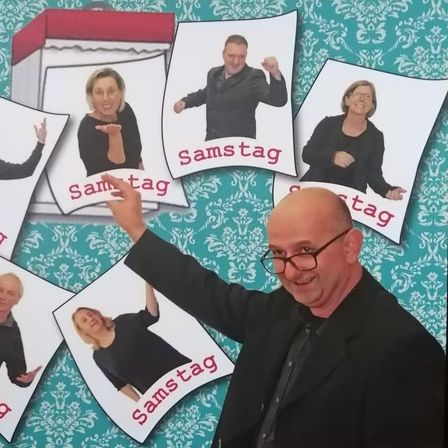 Ein Mann und mehrere Frauen posieren für ein Foto mit dem Wort Samstag darauf. Der Mann lächelt und zeigt auf die Kamera.
