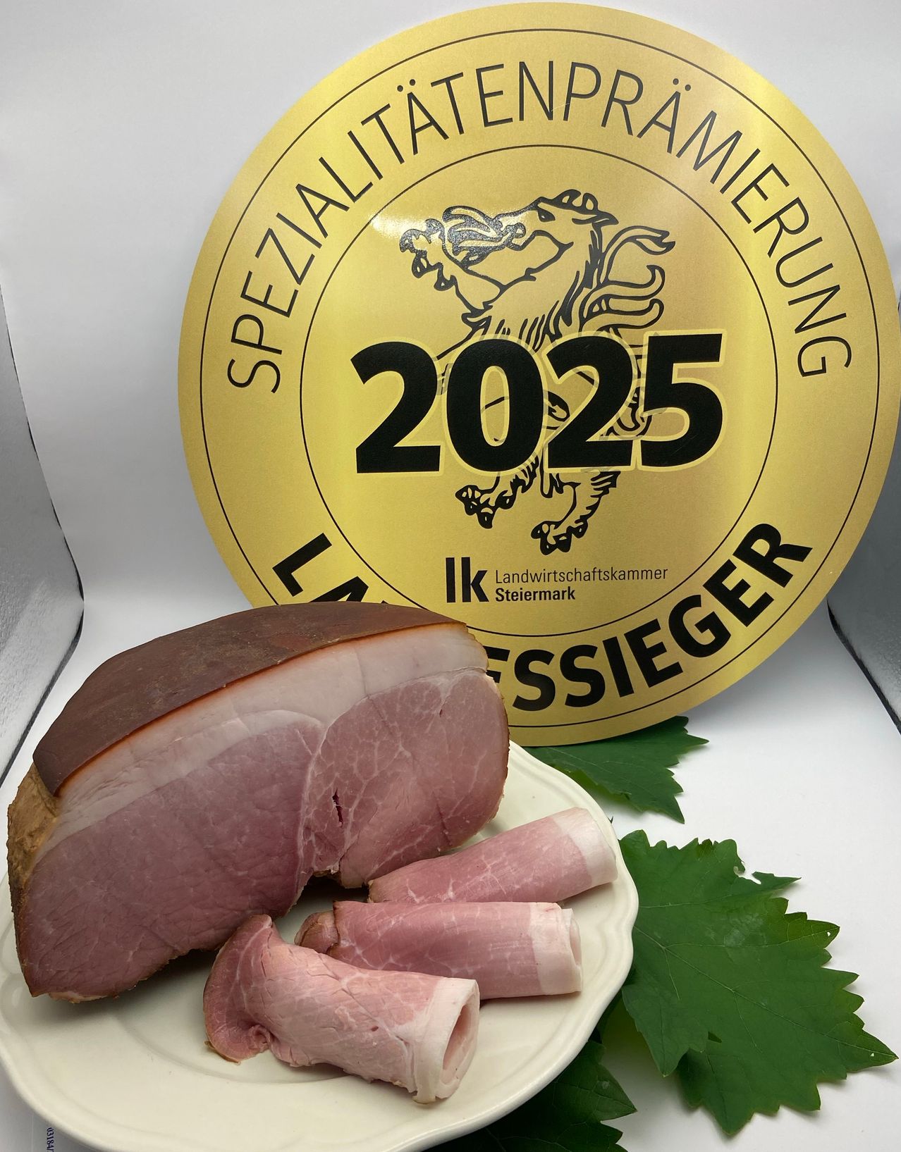 Ein Stück Schinken auf einem Teller mit einem goldenen kreisförmigen Schild, das 'Sonderausstellung 2025' und 'Steiermark' in der Mitte zeigt, zusammen mit einigen Blättern.