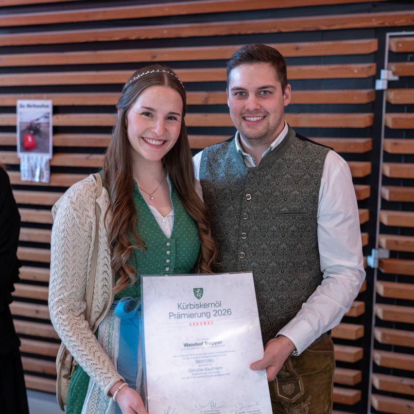 Eine junge Frau und ein Mann stehen lächelnd da und halten ein Zertifikat mit dem Titel 'Kurbiskerl 2026'. Die Frau trägt ein grünes Kleid und einen weißen Cardigan, während der Mann einen grauen Weste und ein weißes Hemd trägt. Sie stehen vor einer Holzwand mit einem Schild und einem roten Objekt.
