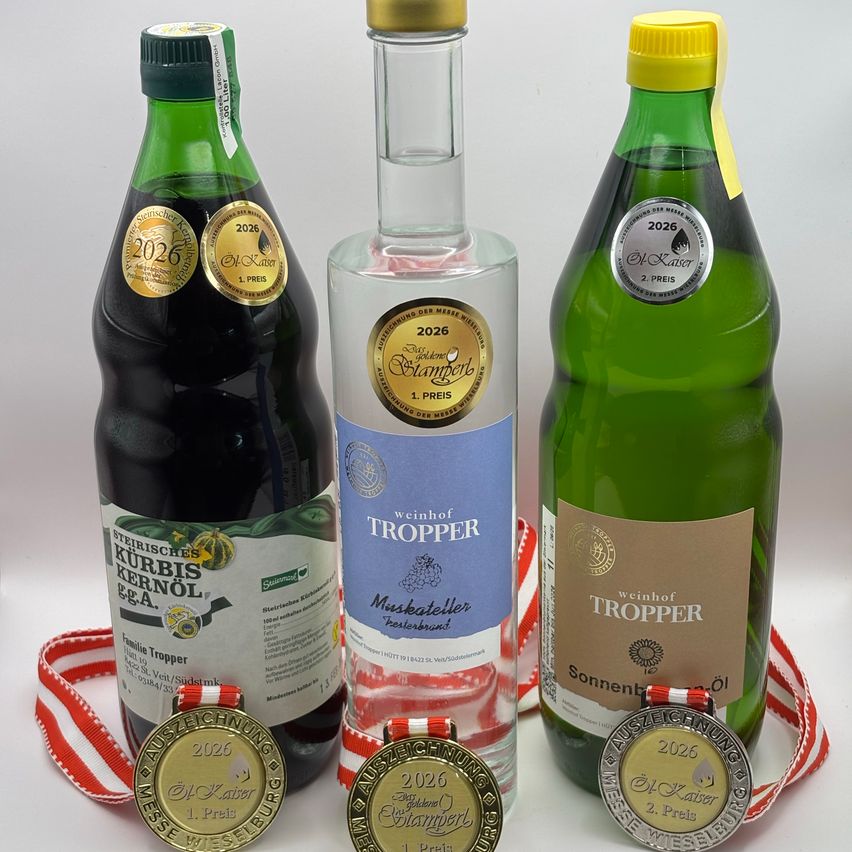 Drei Flaschen Wein und drei Medaillen auf einer weißen Oberfläche. Die Flaschen sind grün mit goldenen Kappen und Etiketten. Eine Flasche hat ein blaues Etikett und zwei Flaschen haben gelbe Kappen. Die Medaillen sind gold und silber mit roten und weißen Bändern.