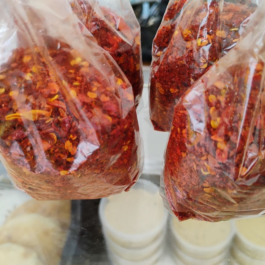 Drei durchsichtige Plastiktüten mit roten und gelben Chiliflocken hängen. Dahinter befinden sich Stapel weißer runder Behälter.